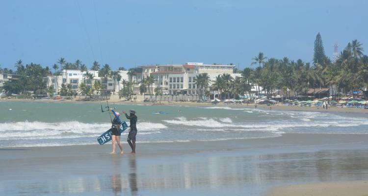 Couple transportant du matériel de kiteboard dans les eaux peu profondes avec les bâtiments de la station balnéaire et les palmiers en arrière-plan.