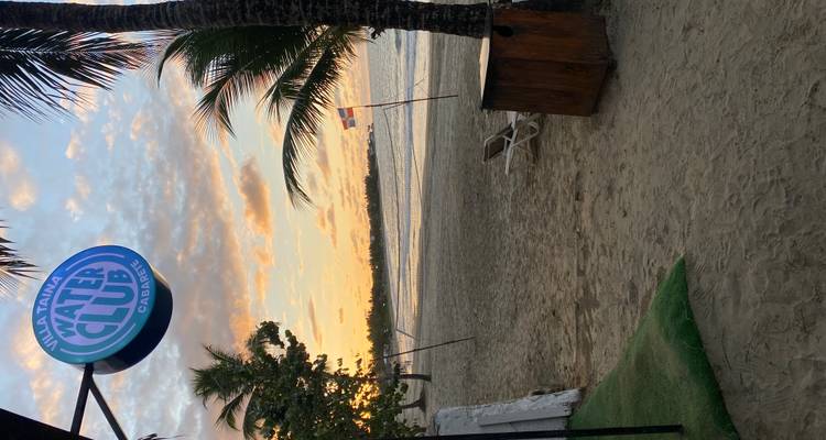 Vue du coucher de soleil sur une plage tranquille encadrée par des palmiers et un panneau indiquant « Villa Taina Water Club Cabarete ».