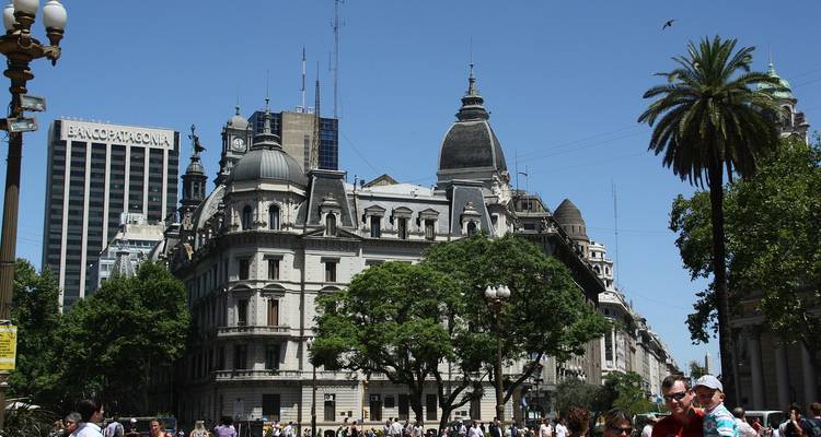 Edificios históricos de estilo europeo rodeando una plaza pública concurrida llena de gente bajo un cielo azul despejado.