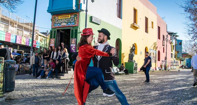 Un hombre y una mujer con trajes vibrantes hacen una pose de tango en la colorida calle de adoquines de Caminito.
