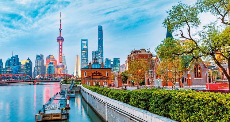 Flussuferpromenade mit üppigem Grünflächenrahmen vor der futuristischen Shanghaier Skyline unter klarem blauem Himmel.