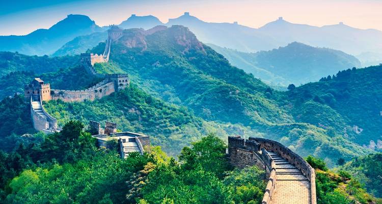Weitreichende Aussicht auf die Chinesische Mauer, die sich über bewaldete Berge unter sanftem Morgennebel schlängelt.