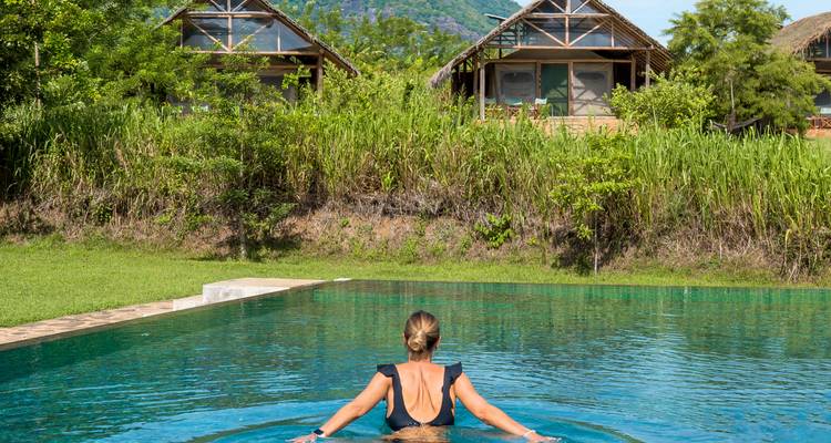 Vrouw in zwemkleding die ontspant in een infinity pool met uitzicht op eco-huisjes en een weelderige tropische heuvel.