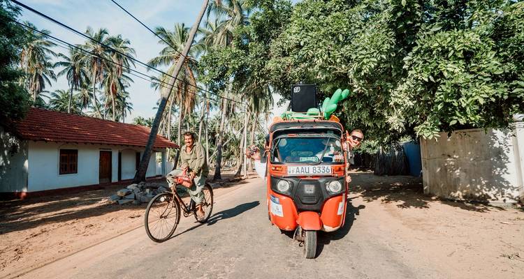 Kleurrijke tuk-tuk met reizigers die poseren naast een lokale fietser op een landelijke Sri Lankaanse weg omgeven door palmbomen.