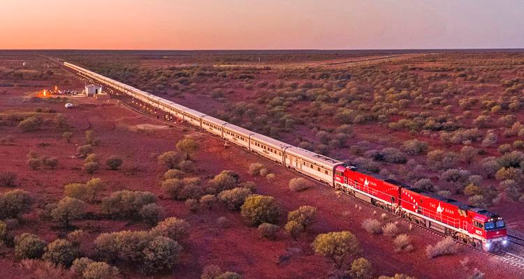 Long train rouge et argent Ghan s'étendant à travers le vaste paysage de l'Outback au lever du soleil avec un feu de camp à proximité.