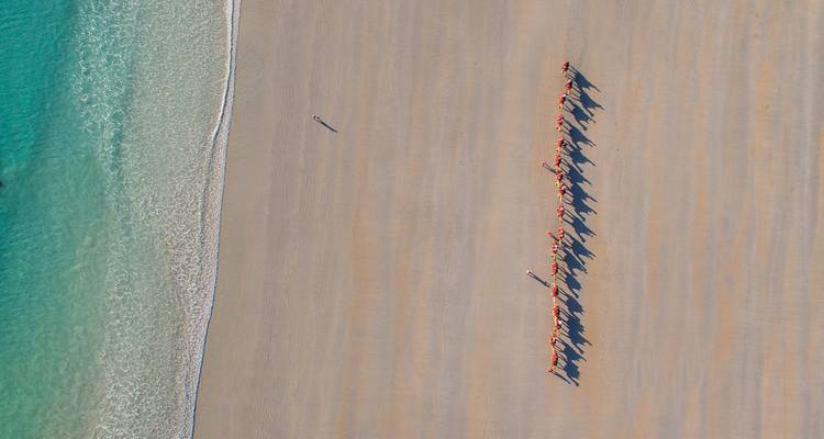 Luchtfoto van een kamelenkaravaan die lange schaduwen werpt op het brede zandstrand van Broome naast turquoise water.