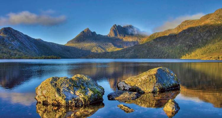 Lac cristallin reflétant les pics escarpés de la Cradle Mountain sous un ciel bleu avec une brume matinale.