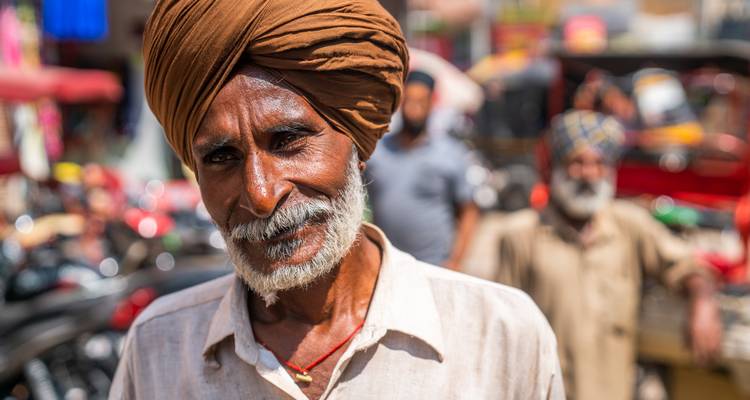 Nahaufnahmeportrait eines lächelnden Sikh-Mannes auf einem belebten indischen Markt mit verschwommenem, farbenfrohem Hintergrund.