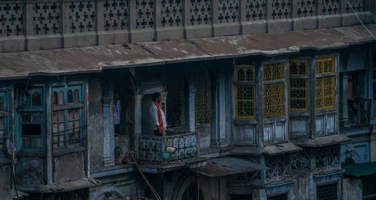 Ein Mann, der auf einem verwitterten Balkon eines alten Gebäudes mit verzierten Holzfenstern in Indien steht.
