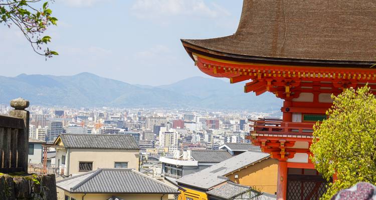 Felgekleurde houten dakranden van een Kyoto-tempel met uitzicht op het stadslandschap en verre bergen.
