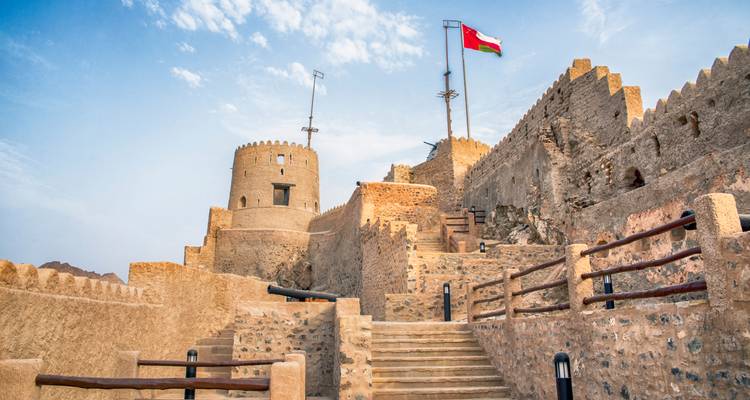 Escalones de piedra que suben hacia un fuerte omaní histórico con la bandera nacional ondeando bajo un cielo azul claro.