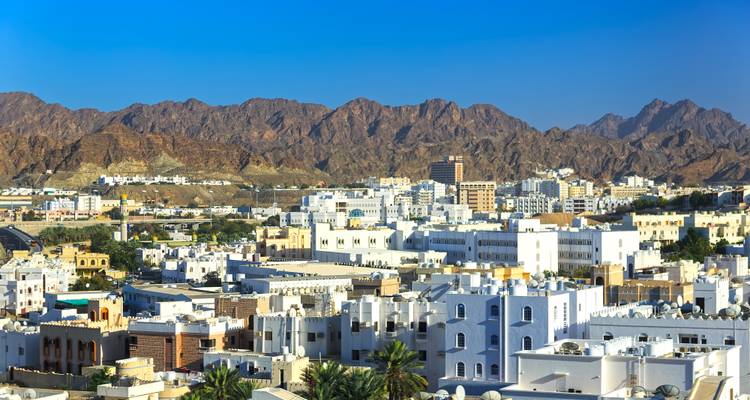 Vista general de los edificios blancos de baja altura de Mascate contra montañas marrones y escarpadas bajo un cielo azul brillante.