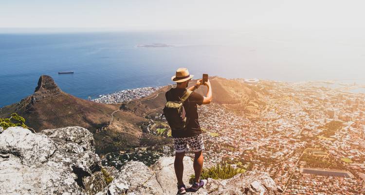 Voyageur avec sac à dos photographiant la vue panoramique de la ville et de la mer depuis un point de vue spectaculaire en falaise.
