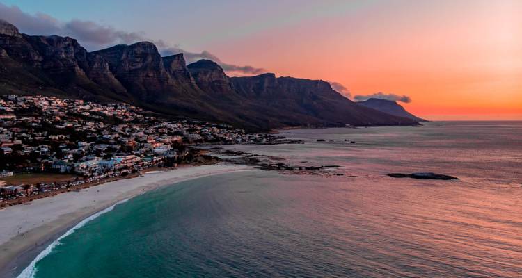 Vue aérienne au coucher du soleil d'une plage sinueuse sous des montagnes escarpées et un ciel aux tons pastel.