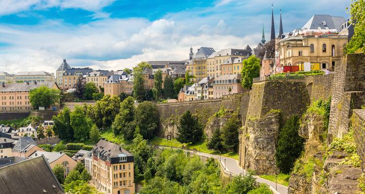 Sun-washed view of Luxembourg City’s cliffs, ramparts and pastel buildings rising above lush greenery.