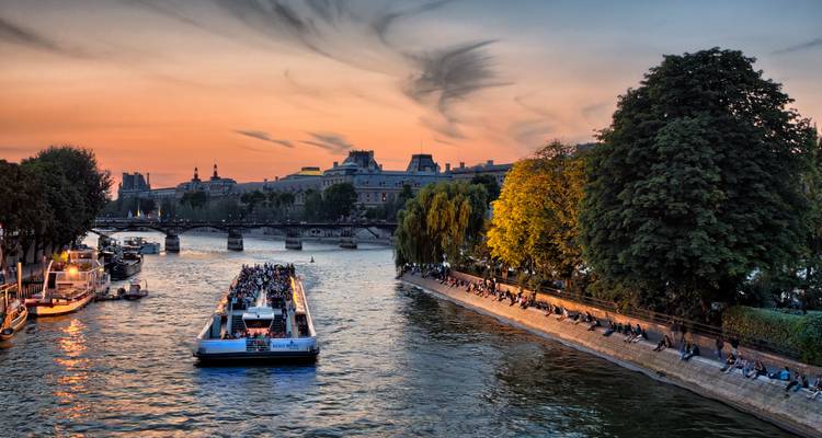 Sightseeing boat cruising a city river at dramatic sunset with silhouettes of bridges and riverside trees.