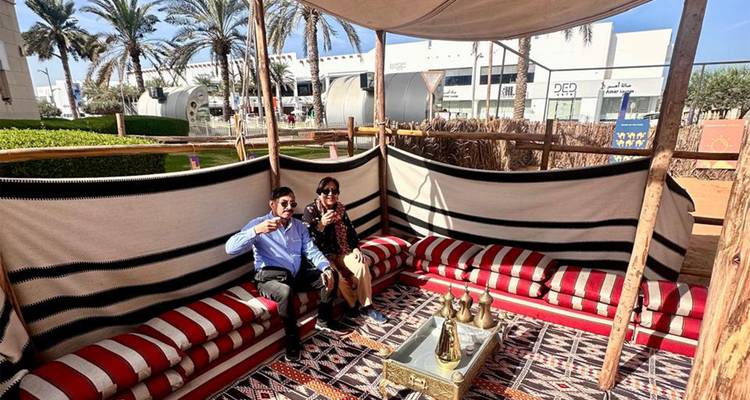 Deux voyageurs savourent un thé arabe sur des coussins à rayures rouges à l'intérieur d'un espace de repos bédouin ombragé.