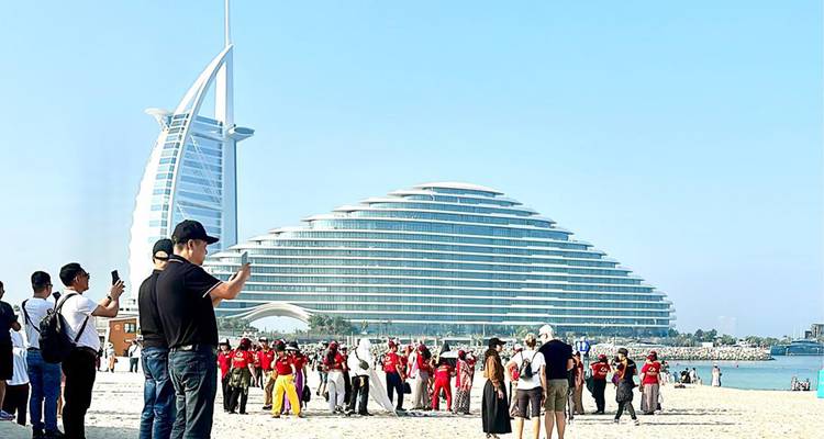 Scène de plage avec des touristes et l'emblématique Burj Al Arab et l'hôtel Jumeirah Beach se détachant sur un ciel bleu dégagé.