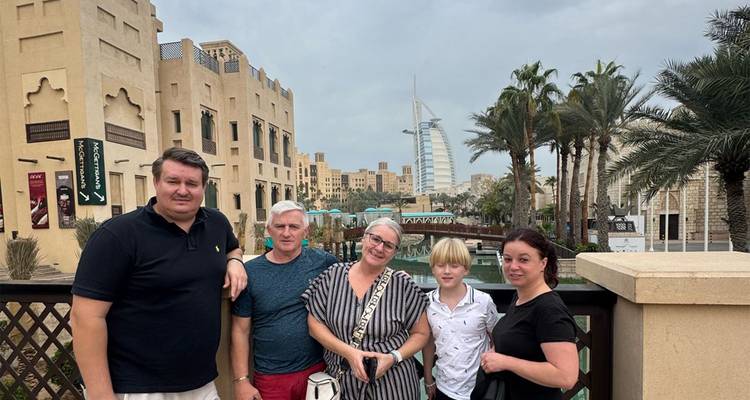 Une famille se tient sur un pont en bois dans une station balnéaire arabique avec des palmiers et la Burj Al Arab encadrant l'arrière-plan.