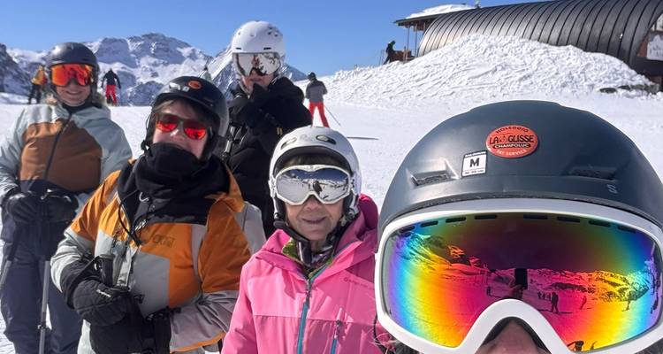 Fröhliche Skifahrer in farbenfroher Ausrüstung machen ein Selfie auf einem sonnigen verschneiten Hang mit Alpengipfeln im Hintergrund.