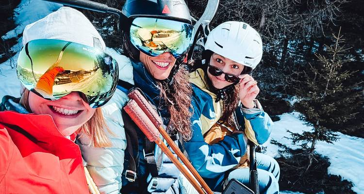 Drei weibliche Freunde auf einem Skilift lächelnd mit reflektierenden Skibrillen und schneebedeckten Bäumen darunter.