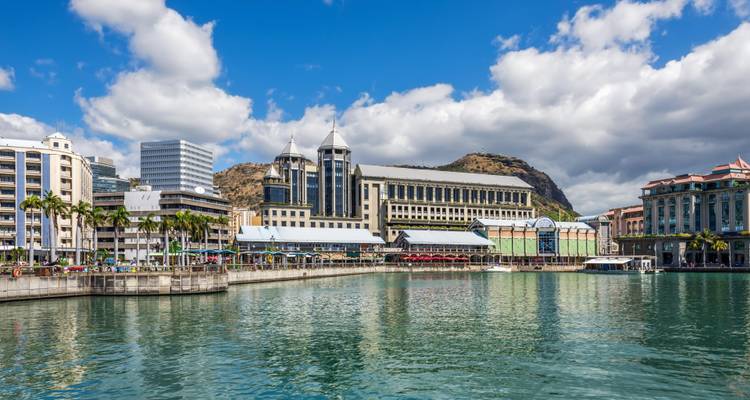 Modern waterfront buildings and mountains reflected in a calm harbor under scattered clouds in Port Louis.