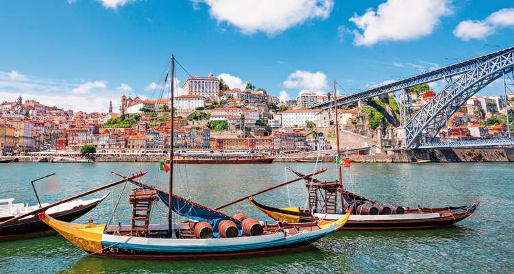 Traditionelle Rabelo-Boote, beladen mit Weinfässern, treiben auf dem Douro-Fluss mit Portos farbenfrohem Ribeira-Viertel und der Dom-Luís-I-Brücke im Hintergrund.
