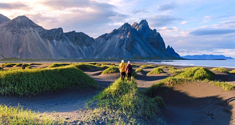 Zwei Wanderer gehen über schwarze Sanddünen, die mit grünem Gras bewachsen sind, während sich die dramatischen gezackten Gipfel des Vestrahorn hinter ihnen erheben.