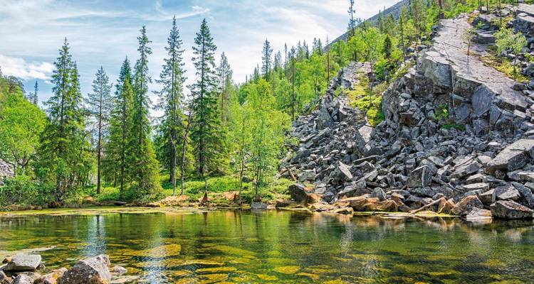 Klarer Waldteich umgeben von Fichtenbäumen und Felshängen unter einem strahlend blauen Himmel.