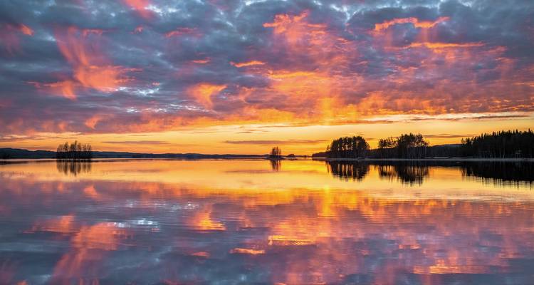 Ein strahlend orangefarbener und rosa Sonnenuntergang spiegelt sich symmetrisch auf einem stillen finnischen See.