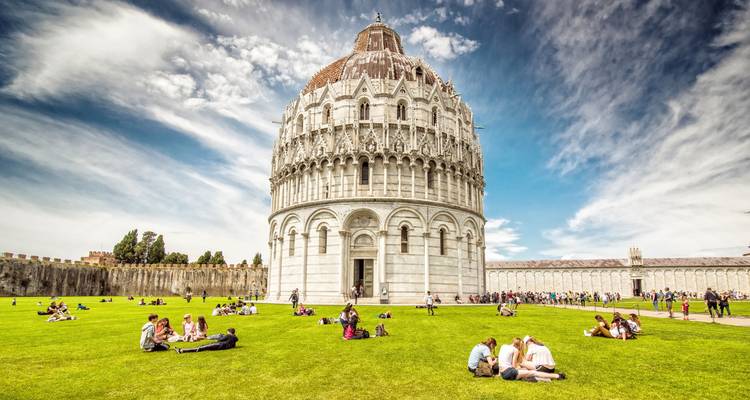 Bezoekers die ontspannen op het groene gazon voor het witte marmeren Doopkapel van Pisa onder een helder blauwe hemel.