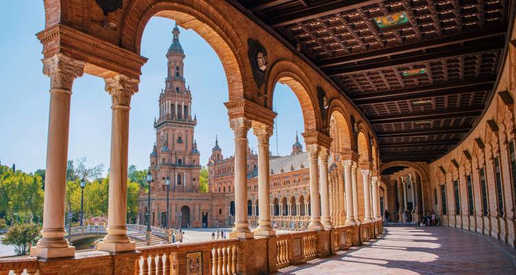 Eine gewölbte Kolonnade umrahmt die kunstvollen Türme der Plaza de España, die in warmem Sevillaner Sonnenlicht erstrahlen.