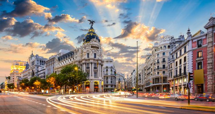 Dramatischer Sonnenuntergang über Madrids Gran Vía mit Lichtspuren des Verkehrs und dem Metropolis-Gebäude im Scheinwerferlicht.