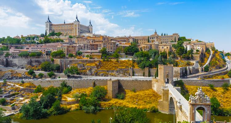 Die historische Stadt Toledo thront auf einem Hügel mit der Alcázar und einer alten Steinbrücke, die den darunter fließenden Fluss überquert.