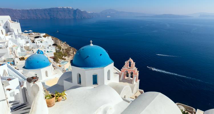 Ikonische weiße Terrassen und blaudomierte Kirchen von Oia mit Blick auf das tiefe Ägäische Meer mit Schnellbooten.