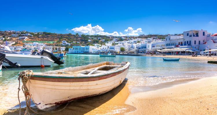 Gestrandetes Holzboot in einer klaren türkisfarbenen Bucht mit weißen Häusern des Hafens von Mykonos.