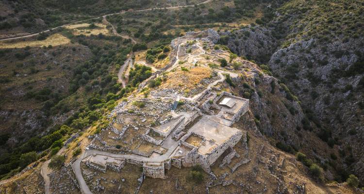 Vue aérienne des ruines d'une ancienne citadelle perchée au sommet d'une colline, entourée de crêtes rocheuses et de pentes parsemées d'oliviers.
