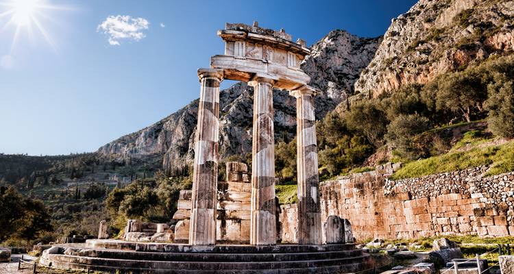 Les colonnes imposantes du Tholos de Delphes se dressent sur fond de montagnes escarpées sous un soleil éclatant.