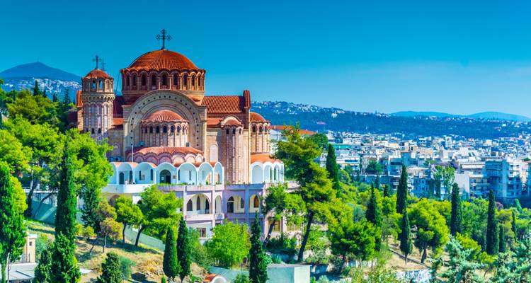 Basilique byzantine entourée de cyprès et skyline moderne de Thessalonique sous un ciel dégagé.