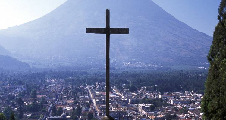 Una cruz en la cima de una colina recortada contra el cielo se alza sobre Antigua y el enorme volcán envuelto en la suave bruma matinal.