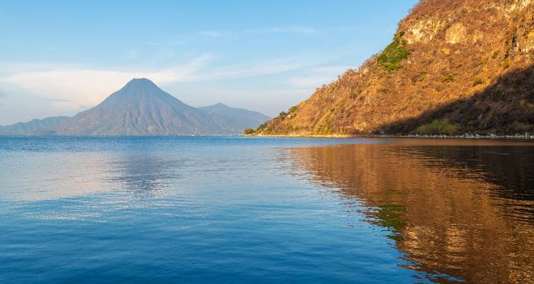 Vista clara a través de un lago tranquilo hacia un volcán cónico y una costa accidentada bajo un cielo azul.