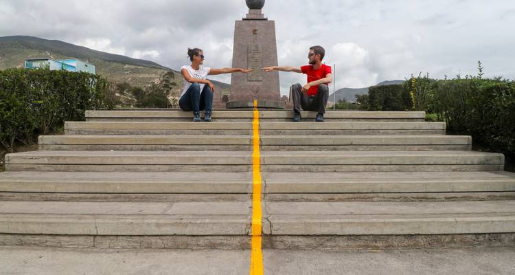 Two travellers sit on opposite sides of the equator line at the Mitad del Mundo monument.