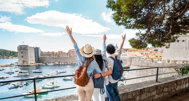 Trois voyageurs joyeux les bras levés surplombant le port de Dubrovnik et ses fortifications.