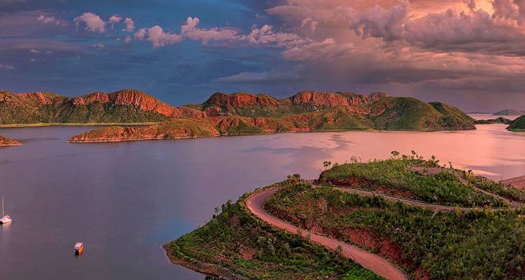 Panoramisches Sonnenuntergangslicht über den Inseln des Lake Argyle und einer gewundenen Straße unter dramatischen Gewitterwolken.