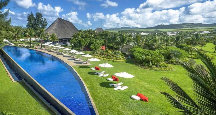 Aerial view of a luxury resort with a long infinity pool, sun-loungers, lush gardens and distant hills in sunshine.