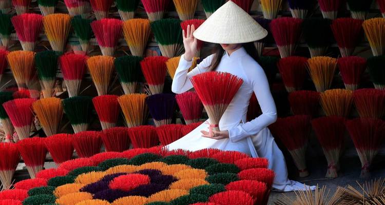 Woman in white ao dai holding red incense sticks, surrounded by colorful arranged bundles, giving a graceful gesture.