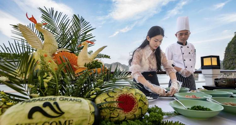 Chef and guest prepare dishes on an open deck with elaborate fruit carvings and karst islands of Halong Bay behind them.
