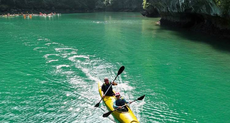 Two kayakers paddle a bright yellow kayak across emerald green water surrounded by limestone cliffs.