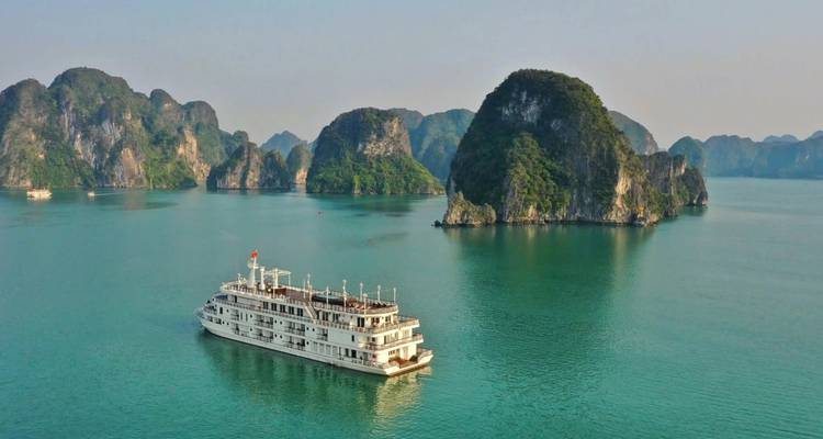 White cruise ship sailing between jungle-covered limestone islands on calm turquoise water in Halong Bay.