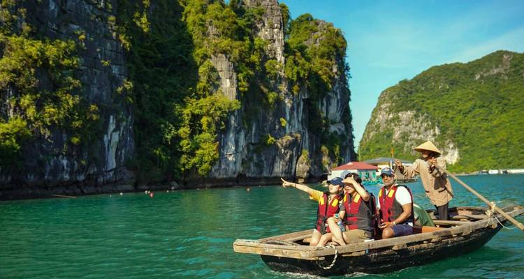 Tourists in life vests ride a traditional wooden rowing boat beneath towering limestone cliffs on blue-green water.